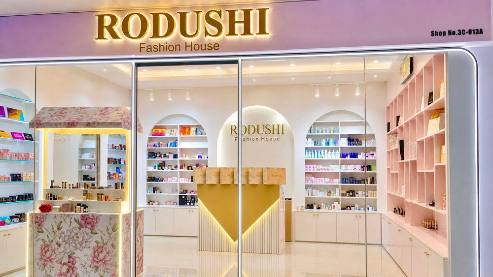 The storefront of RODUSHI Fashion House, featuring a pink and gold sign above glass doors, with a bright, modern interior displaying cosmetics on shelves and a floral-patterned counter.